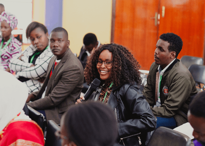A black lady seated in an audience and speaking into a microphone