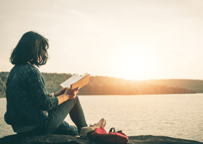 woman reading a book near a water body as the sun is visible at the horizon