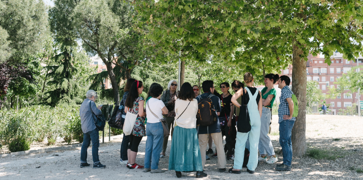 Group of people gathered under a leafy tree in a park, standing in discussion with urban buildings in the background.
